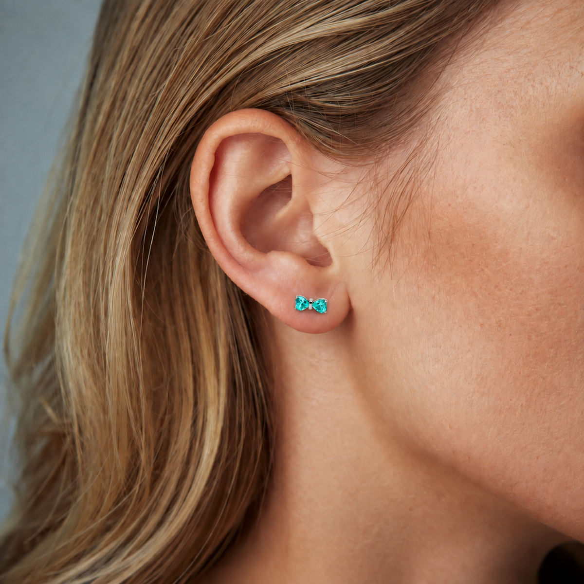 Heart Bow titanium earring with teal heart-shaped CZ stones, worn on the earlobe